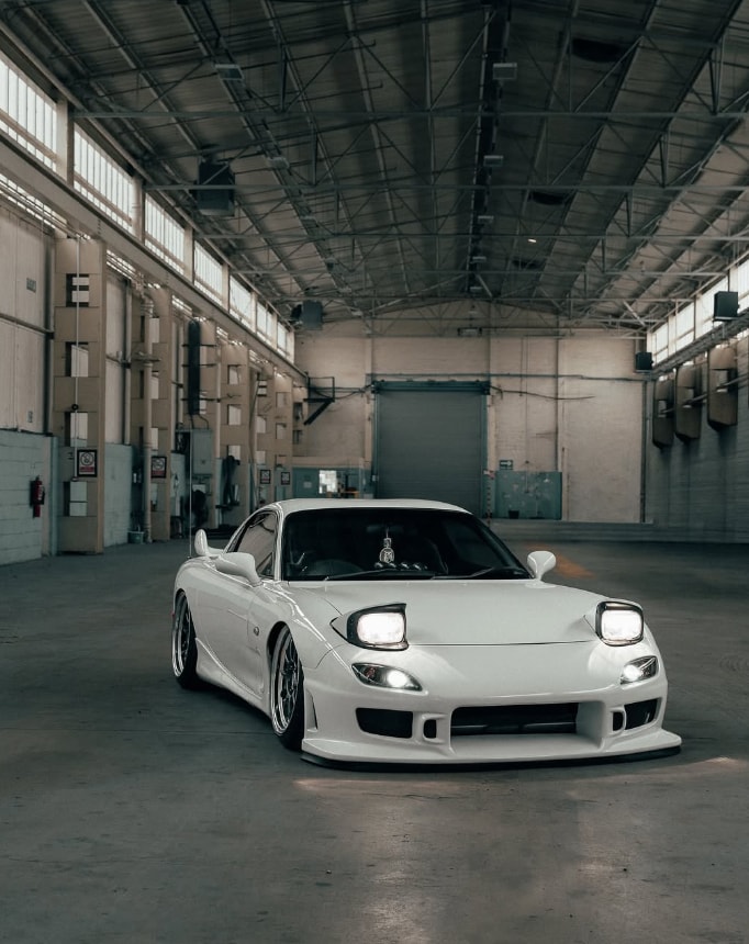 White Mazda RX-7 FD parked low in a large warehouse with pop-up headlights raised